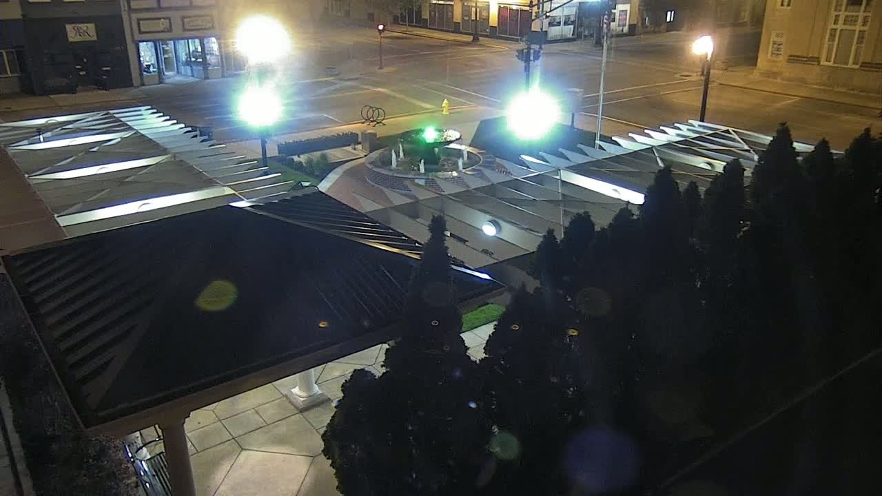 A nighttime aerial view shows a small illuminated fountain surrounded by dark evergreen shrubs and a modern, angular structure with a dark roof.