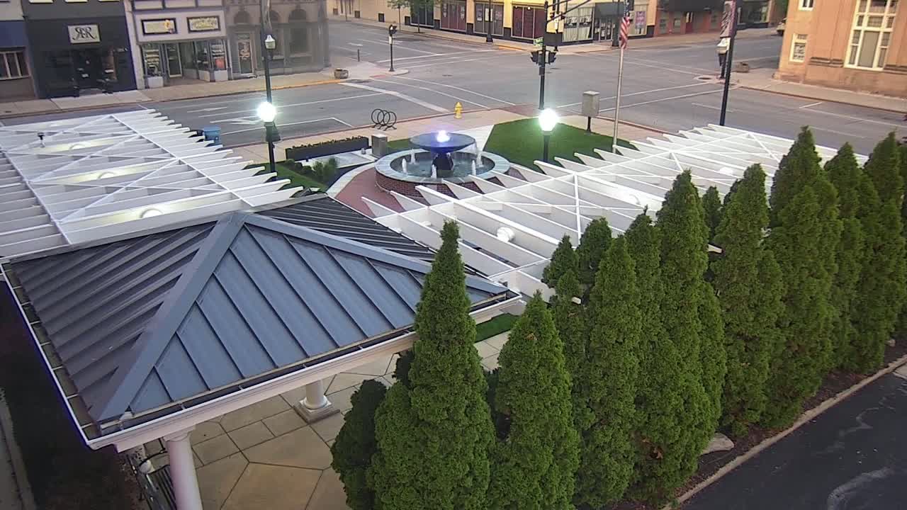 An overhead view shows a dark-blue-roofed pavilion and a white pergola surrounding a fountain in a plaza, all bordered by evergreen trees under a daytime sky.