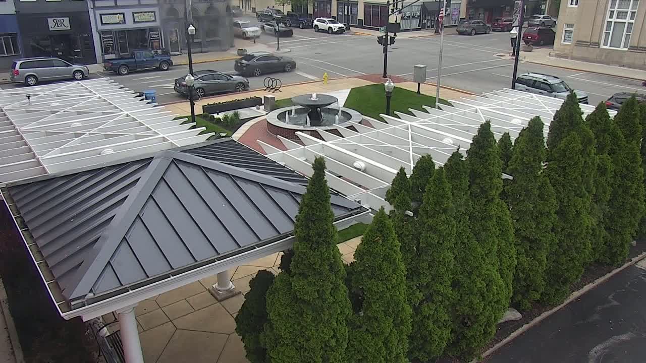 A paved area with a fountain and pergola is surrounded by evergreen trees, with cars parked on a street in the background.