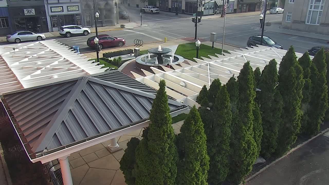 An overhead view shows a town square with a central fountain, surrounded by neatly planted evergreen trees and covered walkways with metal roofs, on a partly sunny day.