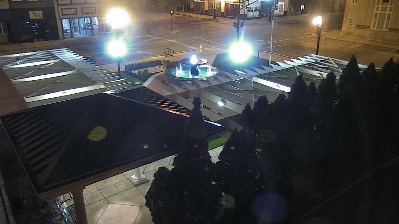 An illuminated fountain is centered in a plaza at night, surrounded by dark evergreen shrubs and a covered structure.