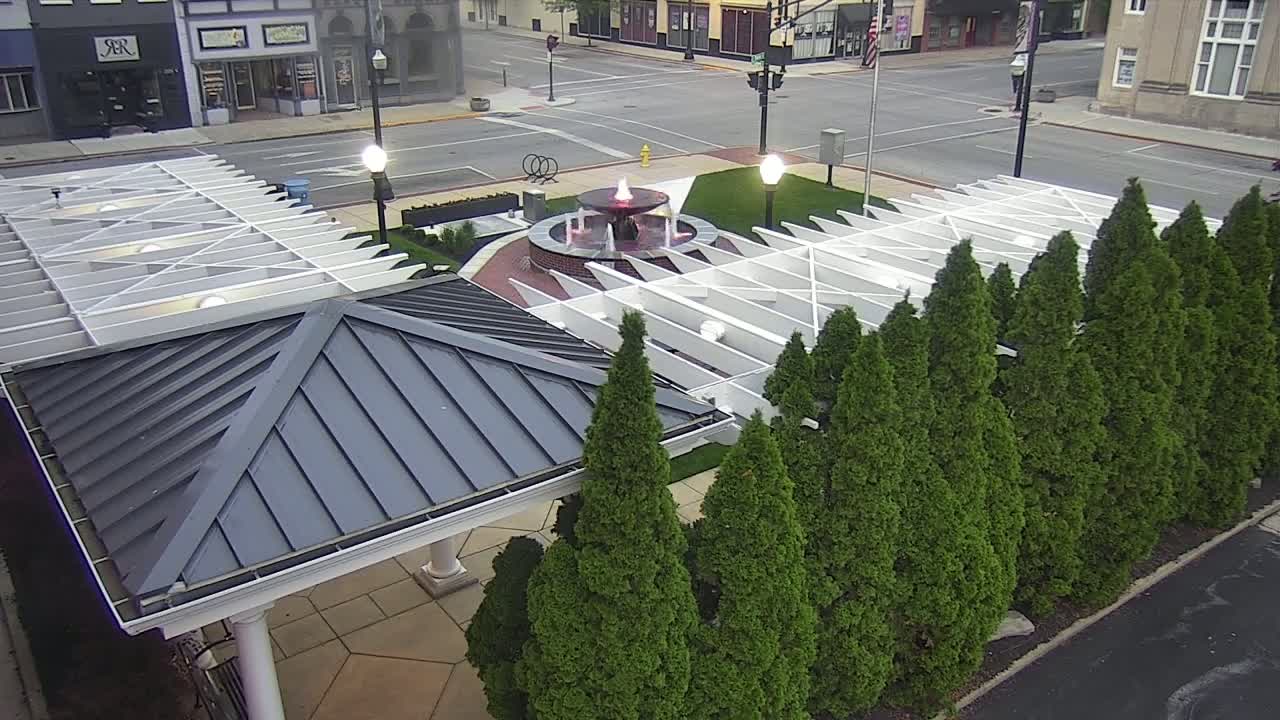 A water fountain is in the center of a town square, surrounded by a row of evergreen trees and a white pergola covering a gray gazebo.