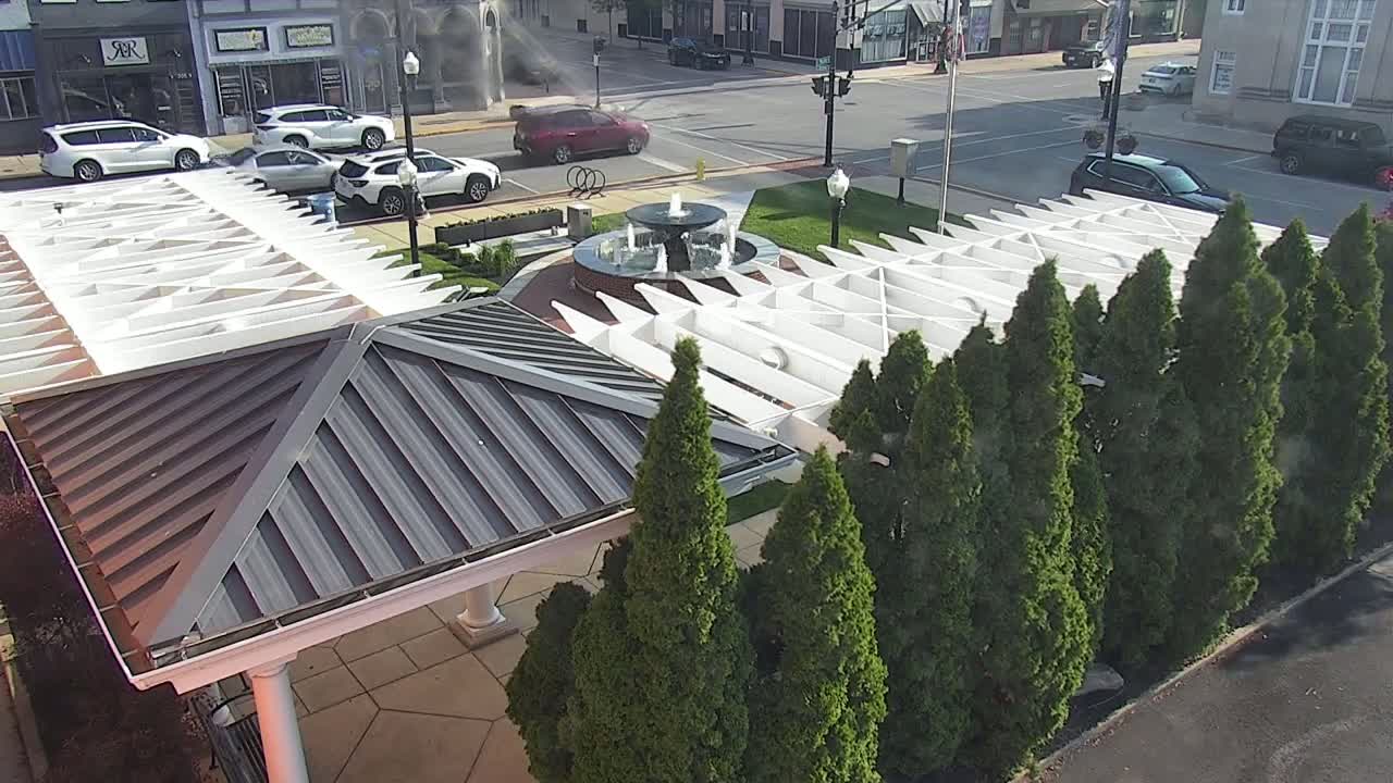 A park scene with a fountain, several cars parked nearby, and a white pergola structure partially shaded by evergreen trees on a sunny day.