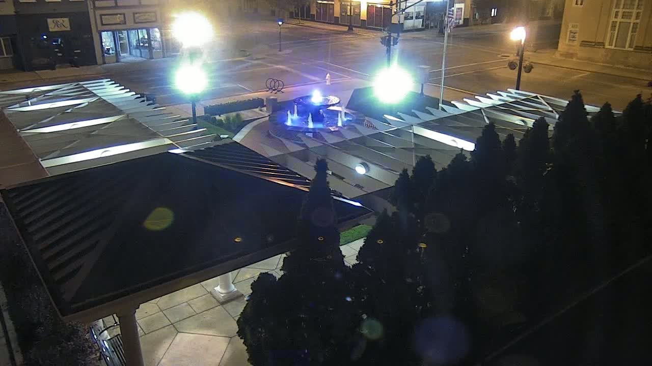 A nighttime view of a city square featuring a lit fountain surrounded by shrubbery and a modern structure with a sloped roof.