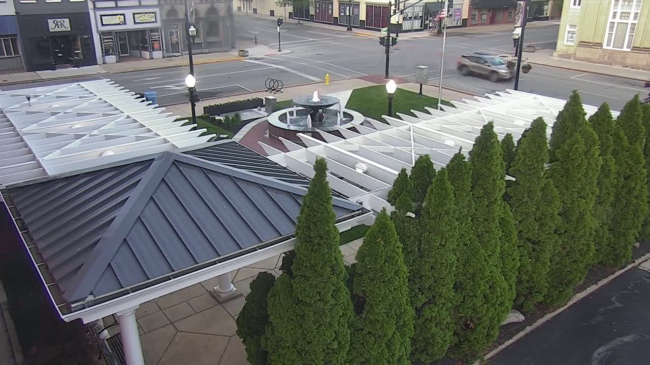 An overhead view shows a paved plaza with a water fountain, a white pergola, and a dark gray gazebo surrounded by evergreen trees, under a mostly clear sky.