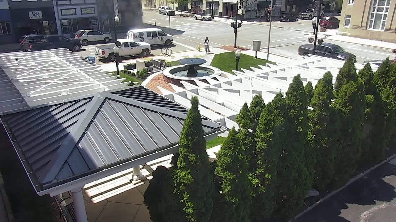 A modern, angular structure with a dark gray roof and a white trellis-like top, partially shaded by a row of evergreen trees, surrounds a central fountain in a sunny city plaza.