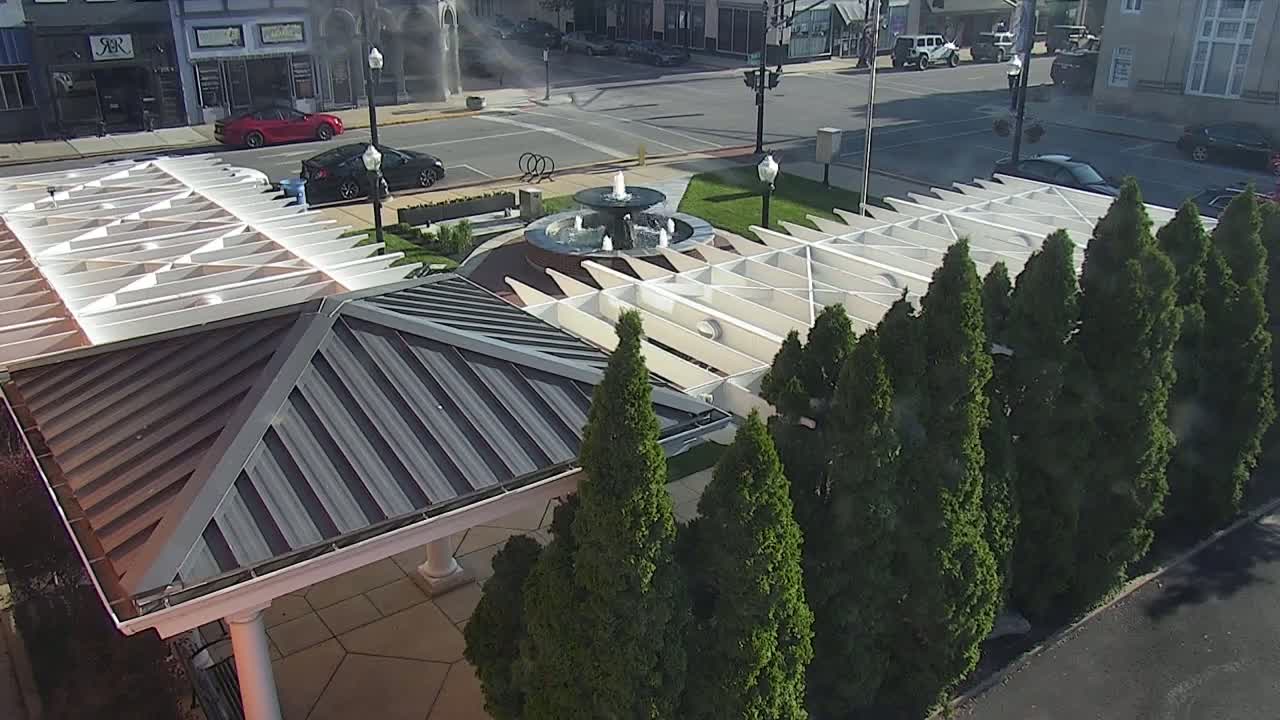 A gazebo with a metal roof sits beside a fountain surrounded by a row of evergreen trees, all in a sunny area.