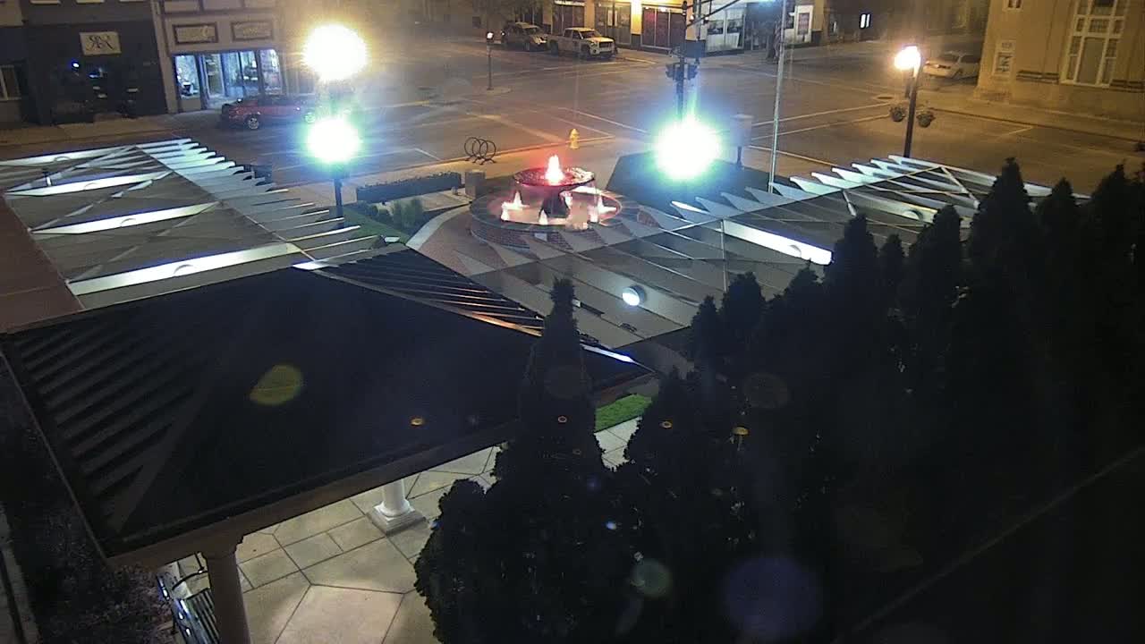A nighttime, overhead view of a lit fountain in a plaza surrounded by dark evergreen shrubs and a modern, dark-colored roof structure.