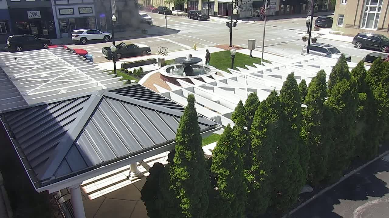 A view from above shows a paved area with a fountain, a row of evergreen trees, and a gray metal-roofed structure; cars are parked on the street beyond.