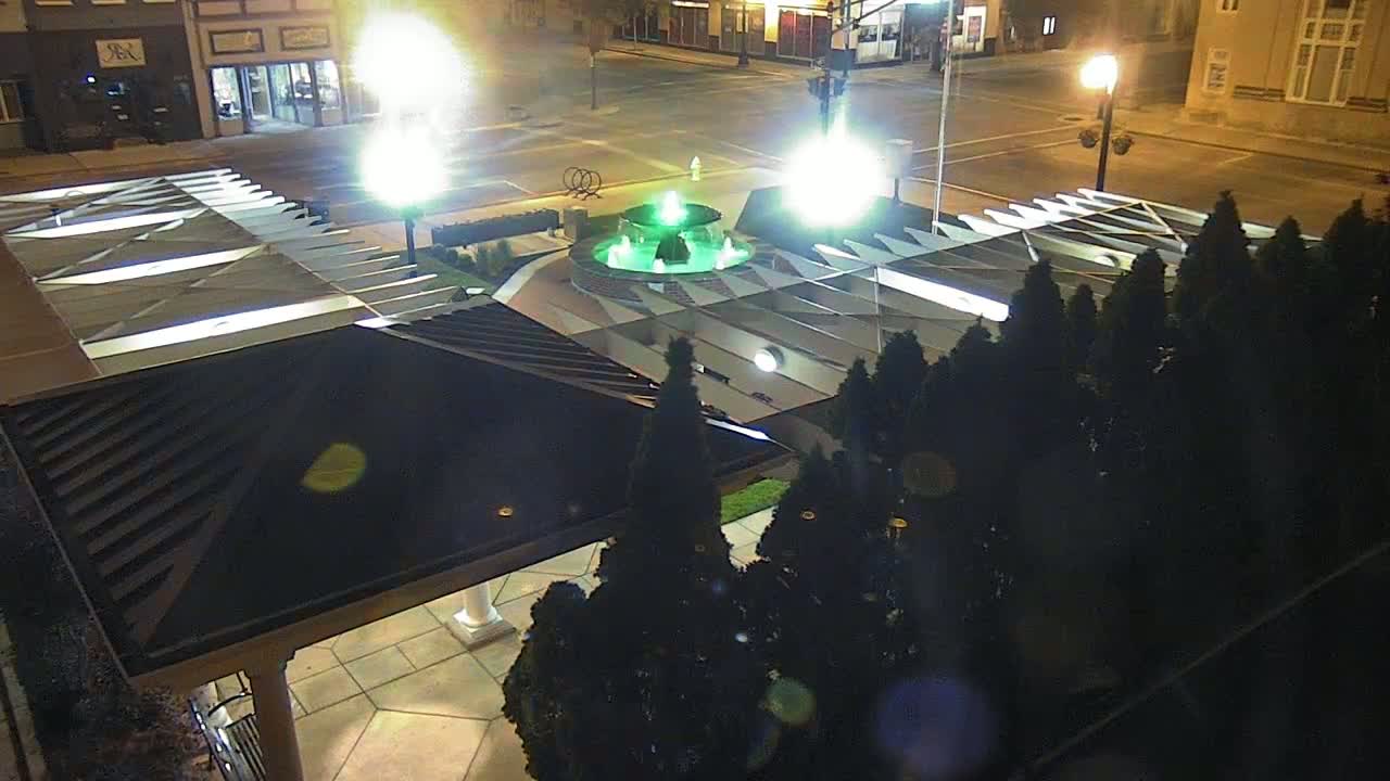 At night, a brightly lit green fountain is centered in a plaza surrounded by dark evergreen trees and a partially visible building with a dark, angled roof.
