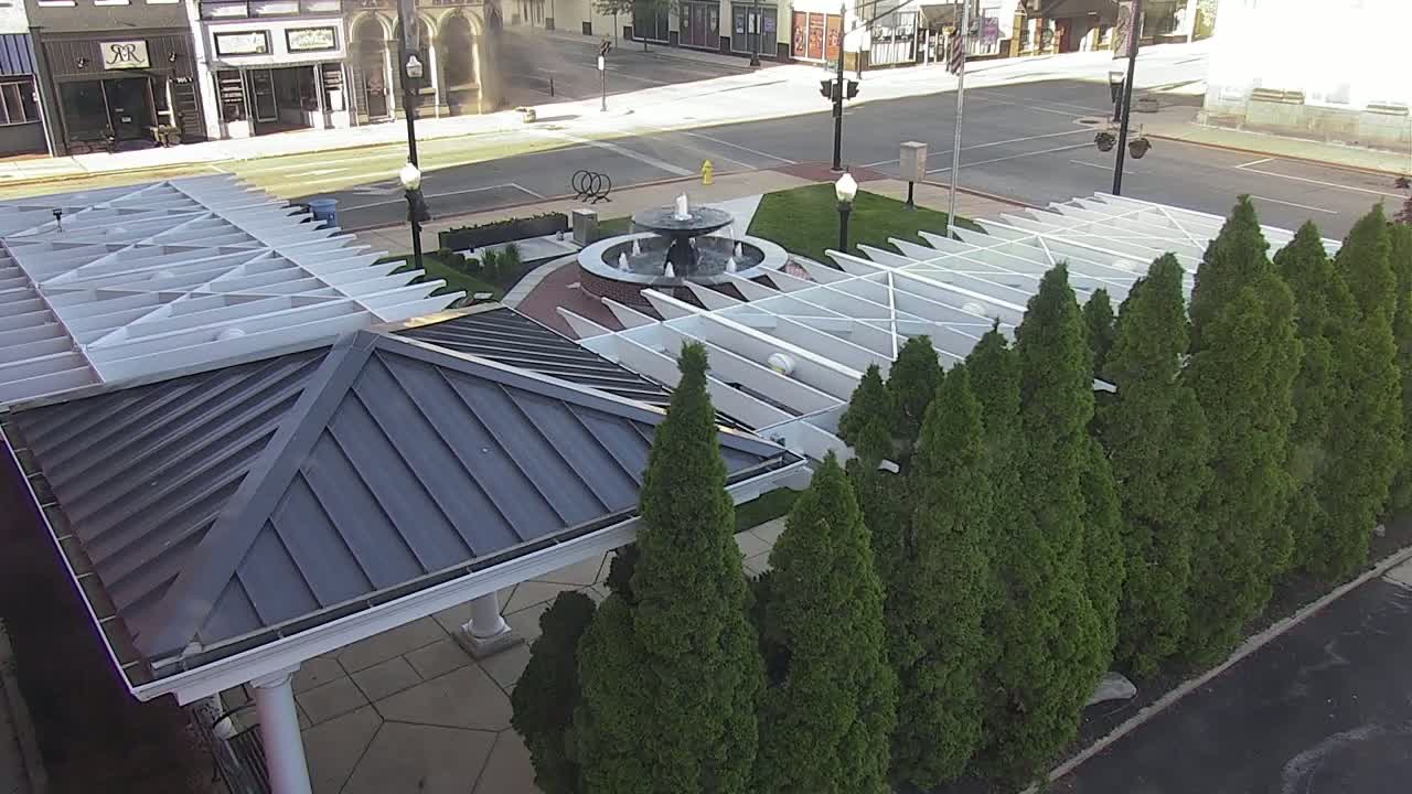 An overhead view shows a paved plaza with a central fountain, surrounded by a row of evergreen trees and covered walkways with modern architectural designs.