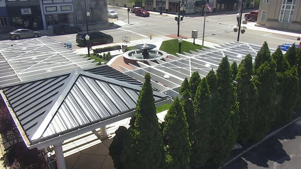 An overhead view of a modern plaza with a central fountain, surrounded by evergreen trees and a metal-roofed pavilion on a sunny day.