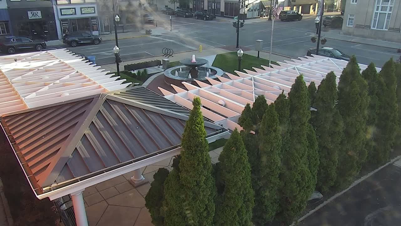 An overhead view shows a water fountain in a paved plaza surrounded by evergreen trees and a metal-roofed pergola on a partly cloudy day.