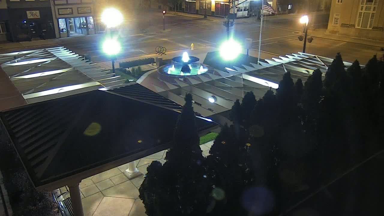 A nighttime aerial view of a fountain in a plaza, surrounded by trees and illuminated by streetlights.