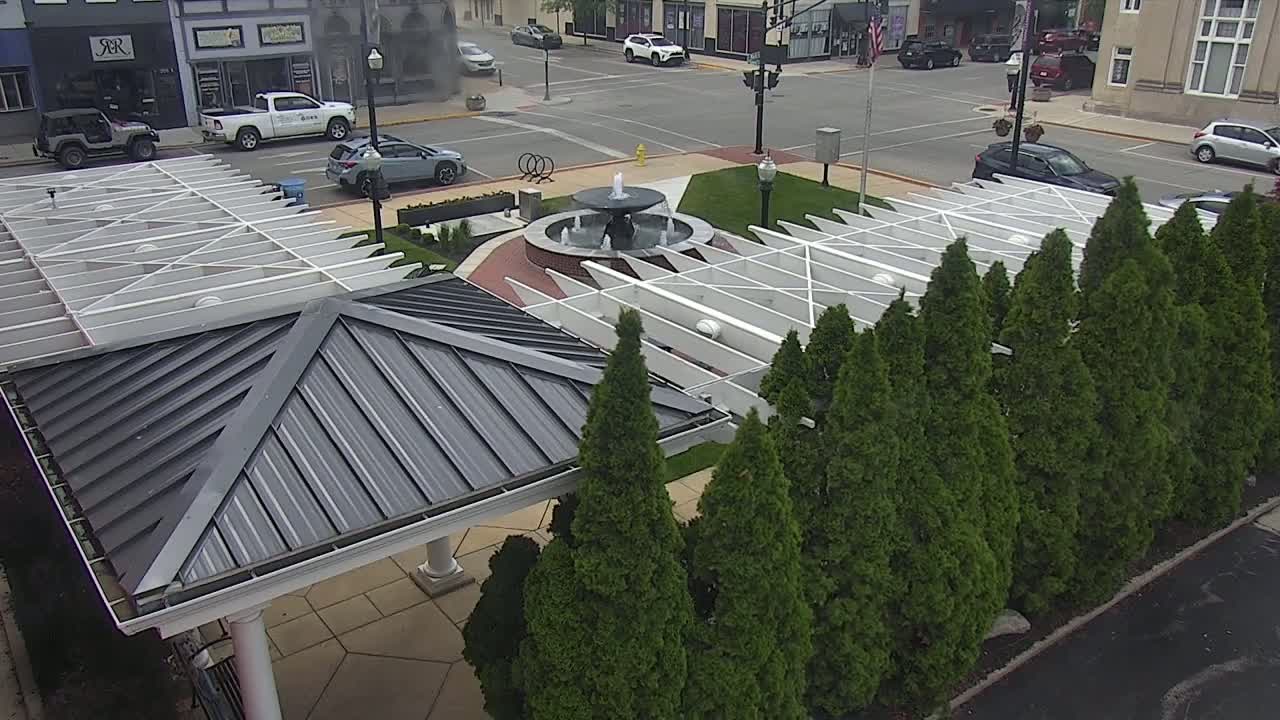 A paved area with a fountain features a white pergola structure partially shaded by a dark-grey roofed pavilion, surrounded by evergreen trees, next to a street with parked cars.