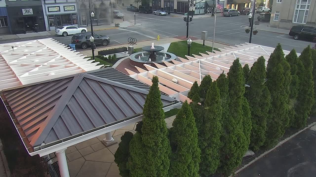 An overhead view of a park-like setting with a fountain at its center, surrounded by evergreen trees and a modern metal and wood pergola structure.