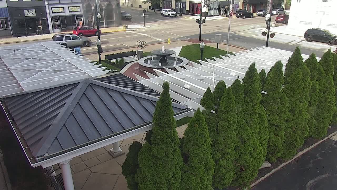 A paved plaza with a central fountain, surrounded by a row of evergreen trees and a white pergola-covered structure with a dark gray metal roof.