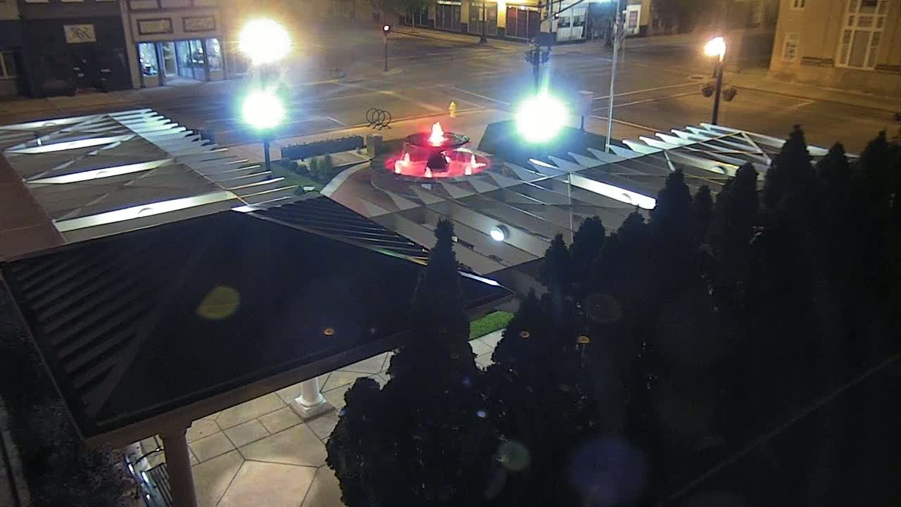 An illuminated fountain with red water is the centerpiece of a plaza at night, surrounded by dark evergreen shrubs and brightly lit buildings.