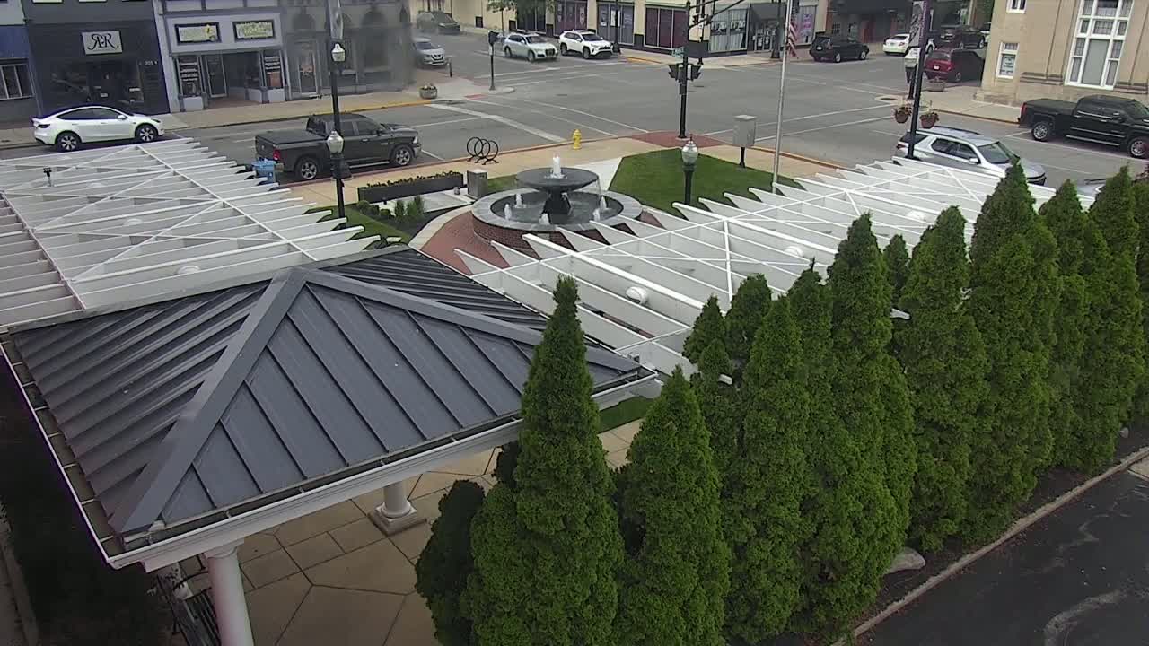An overhead view shows a town square with a fountain, white pergola structures, and a row of evergreen trees on a partly cloudy day.