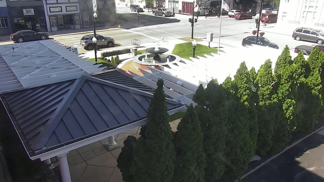 A high-angle view shows a paved area with a fountain in a small park, surrounded by evergreen shrubs and a partially visible building with a dark gray metal roof and a white pergola.