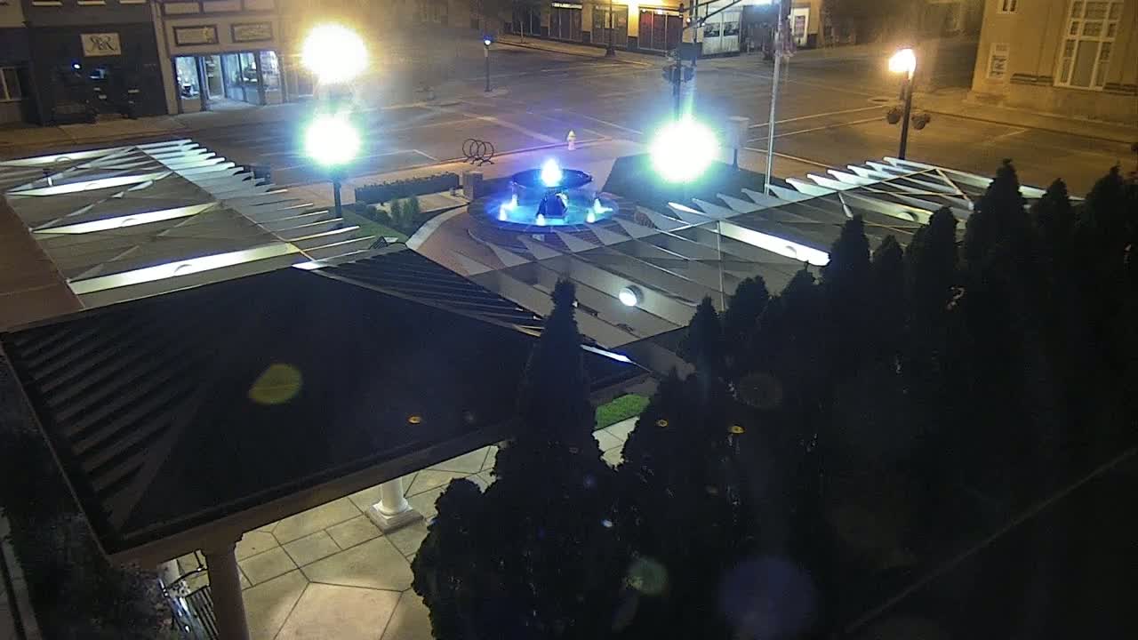 An illuminated fountain is centered in a plaza at night, surrounded by dark evergreen shrubs and a modern, partially covered structure.