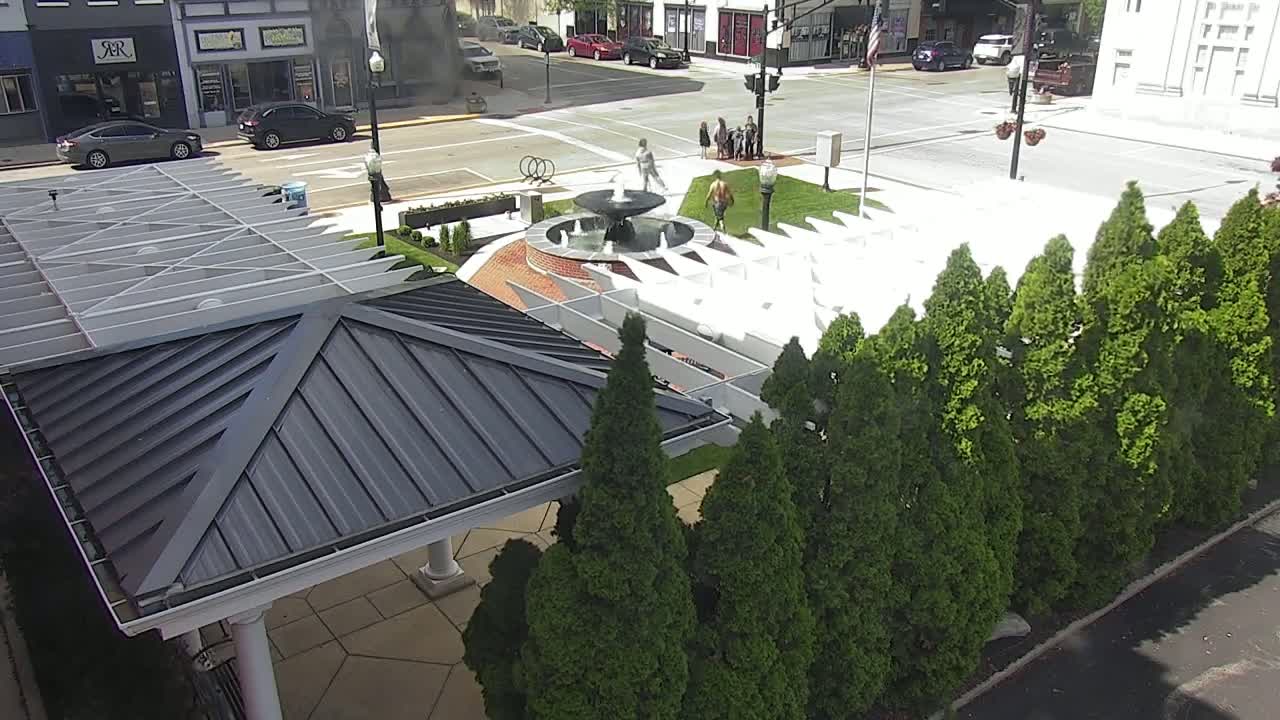 An aerial view shows a paved town square with a fountain, several people, and a row of evergreen trees beside a dark-grey-roofed gazebo under a sunny sky.