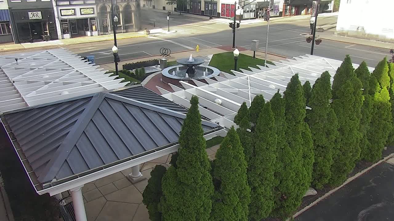 An aerial view shows a gazebo with a dark gray metal roof next to a water fountain in a plaza surrounded by evergreen trees on a sunny day.