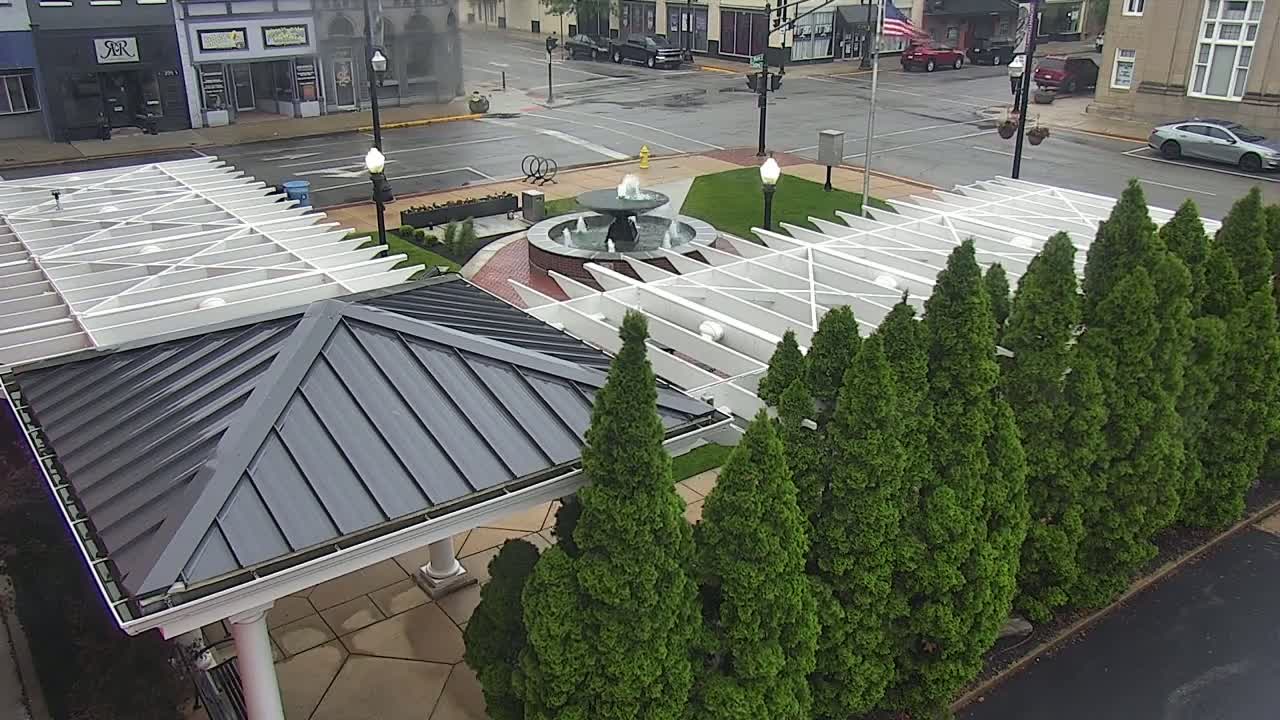 A water fountain in a town square is surrounded by a row of evergreen trees and a white pergola under a light rain.