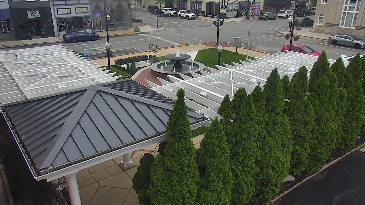 An overhead view shows a town square with a fountain, surrounded by a row of evergreen trees and a white pergola with a dark gray roof.