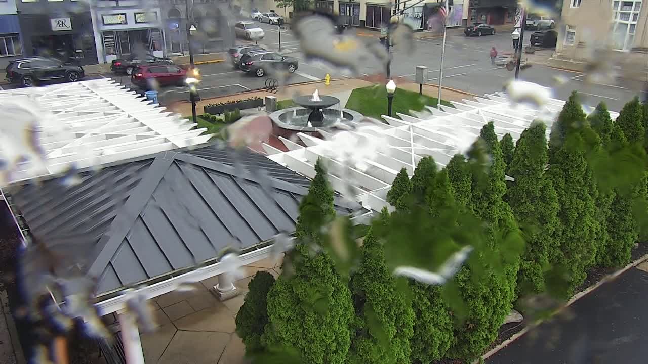 A rain-covered view of a gazebo with a dark gray roof and white pergola over a fountain in a courtyard surrounded by evergreen shrubs, with cars and buildings visible in the background.