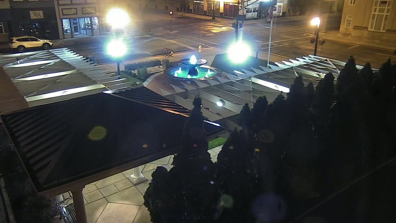 An illuminated fountain is centered in a plaza at night, surrounded by dark evergreen shrubs and a partially visible covered structure.