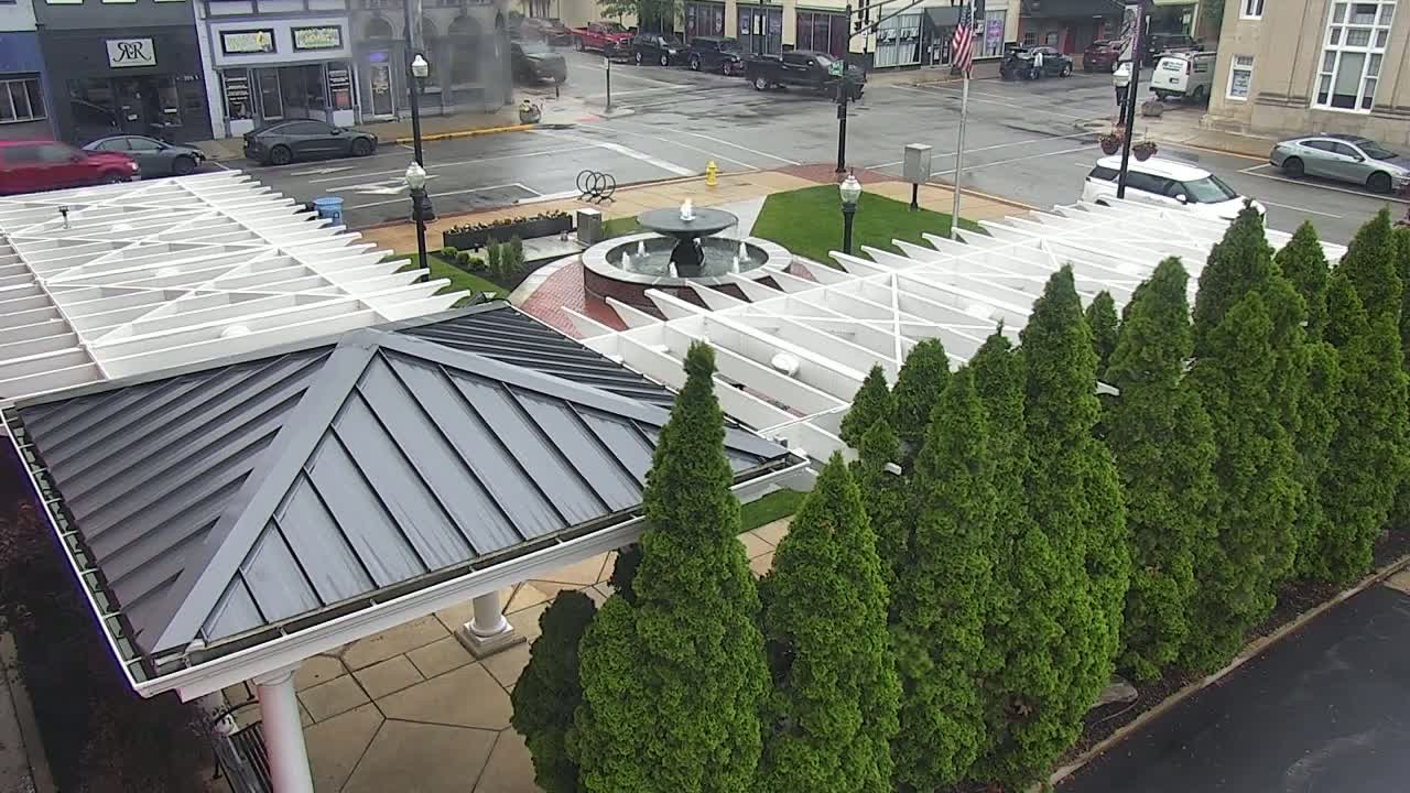 A wet town square features a central fountain surrounded by a row of evergreen trees and a white pergola with a dark gray roof.