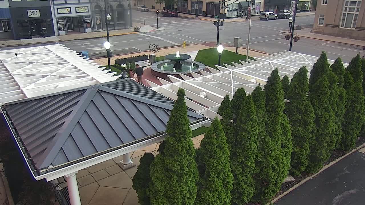 A paved plaza with a central fountain, surrounded by a row of evergreen trees and covered structures with white and gray roofs, on a sunny day.