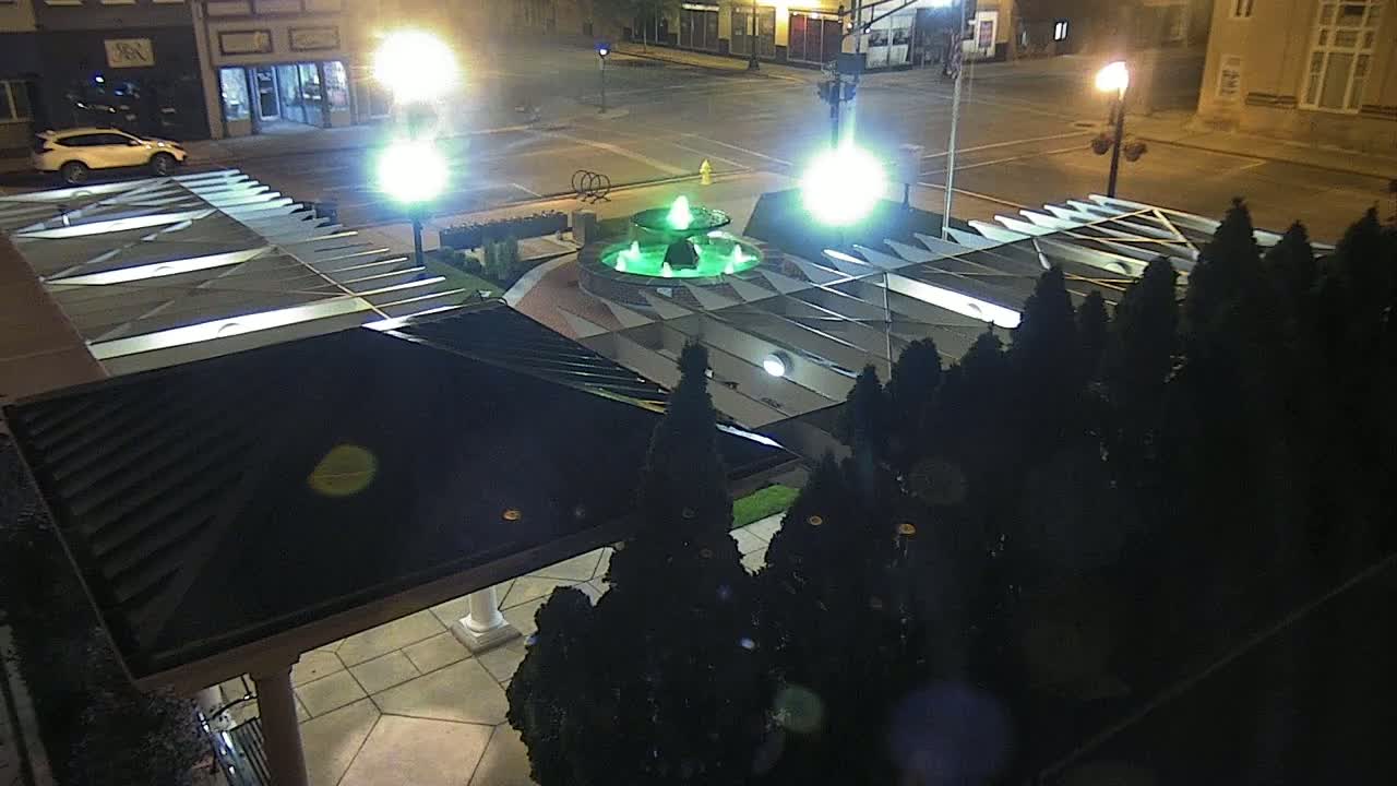 A green-lit fountain is centered in a town square at night, surrounded by dark evergreen shrubs and a covered structure.