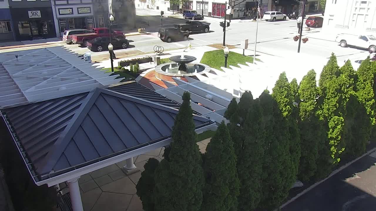 A dark-roofed gazebo sits beside a row of evergreen trees near a fountain in a sunny town square.