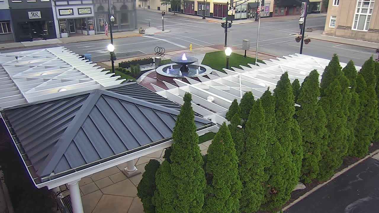 A water fountain is situated in a town square, surrounded by a white pergola and rows of evergreen trees under a partly cloudy sky.