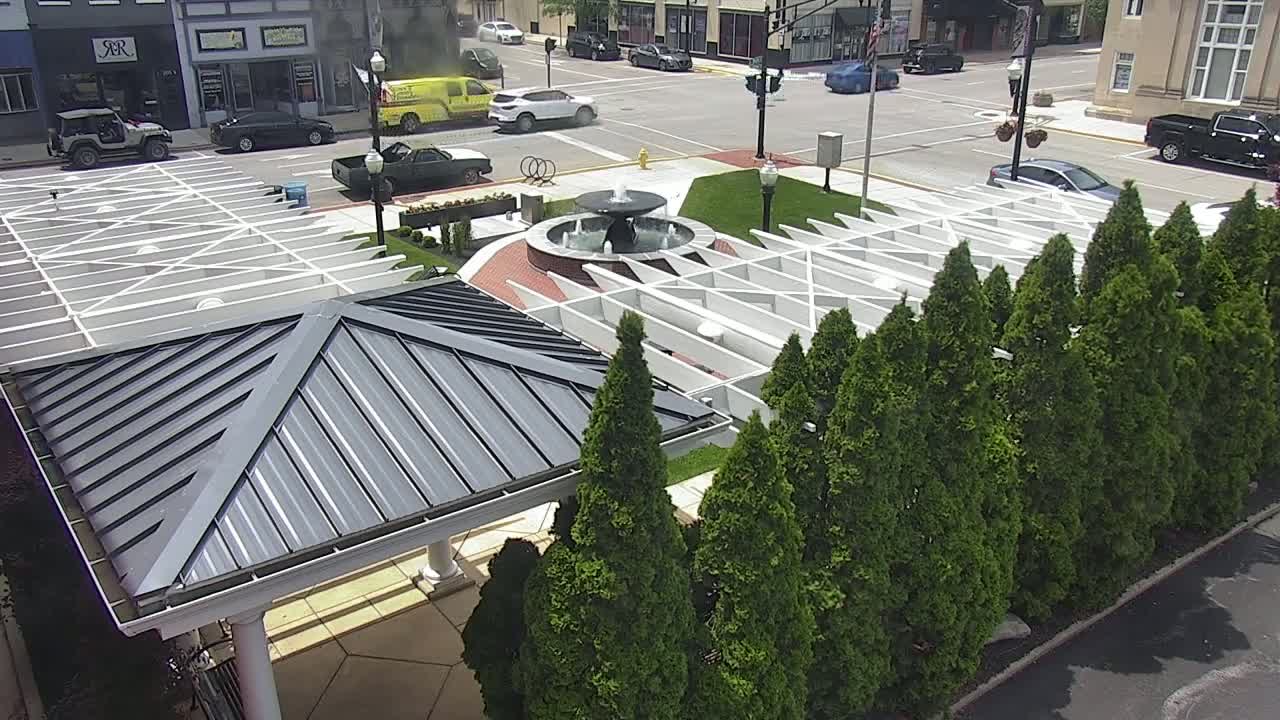 An aerial view shows a town square with a fountain, a white pergola structure, and a row of evergreen trees, all under a sunny sky.