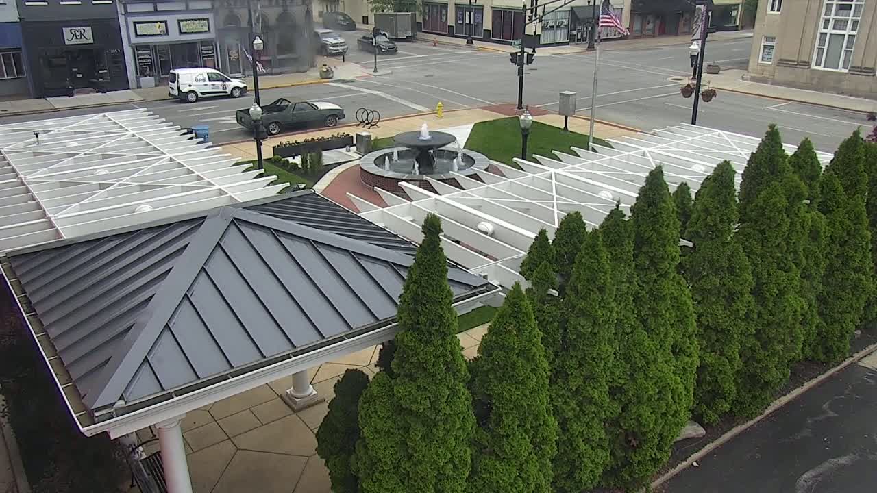 An overhead view shows a town square with a fountain, a white pergola structure, and a row of evergreen trees under an overcast sky.