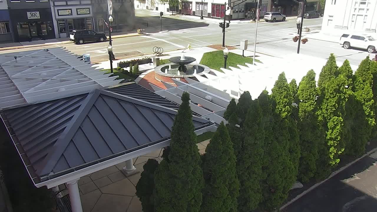 A dark-roofed gazebo sits beside a small fountain and a row of evergreen trees in a sunny plaza, with a street and buildings visible in the background.