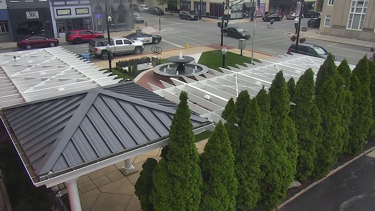 A gray-roofed gazebo with a white pergola sits beside a row of evergreen trees and a fountain in a town square.