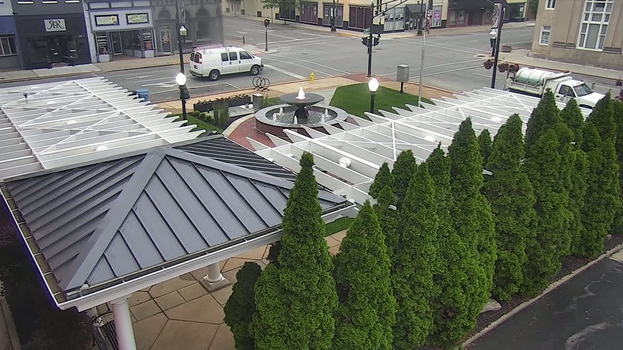A gray-roofed gazebo sits beside a row of evergreen trees near a central fountain in a town square, under an overcast sky.