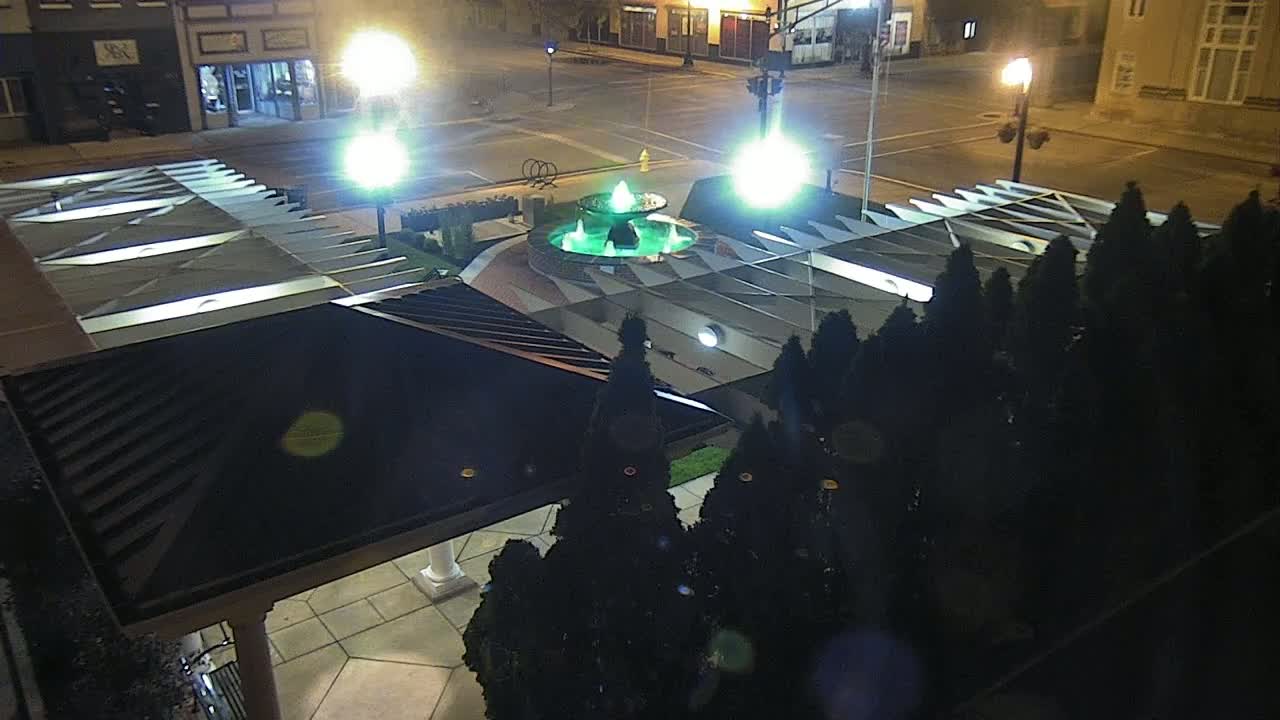 A nighttime, overhead view shows a green fountain in a plaza surrounded by dark evergreen shrubs, illuminated by bright streetlights.