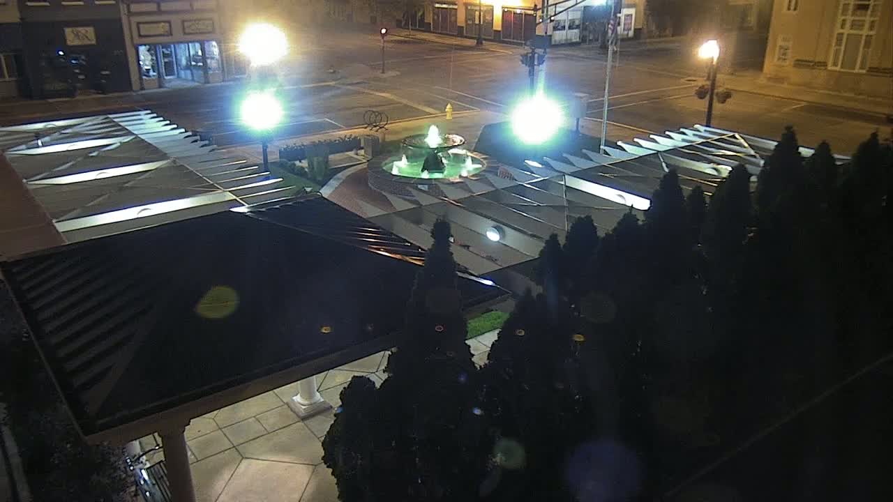 An illuminated fountain is centered in a plaza at night, surrounded by dark evergreen shrubs and a covered walkway.