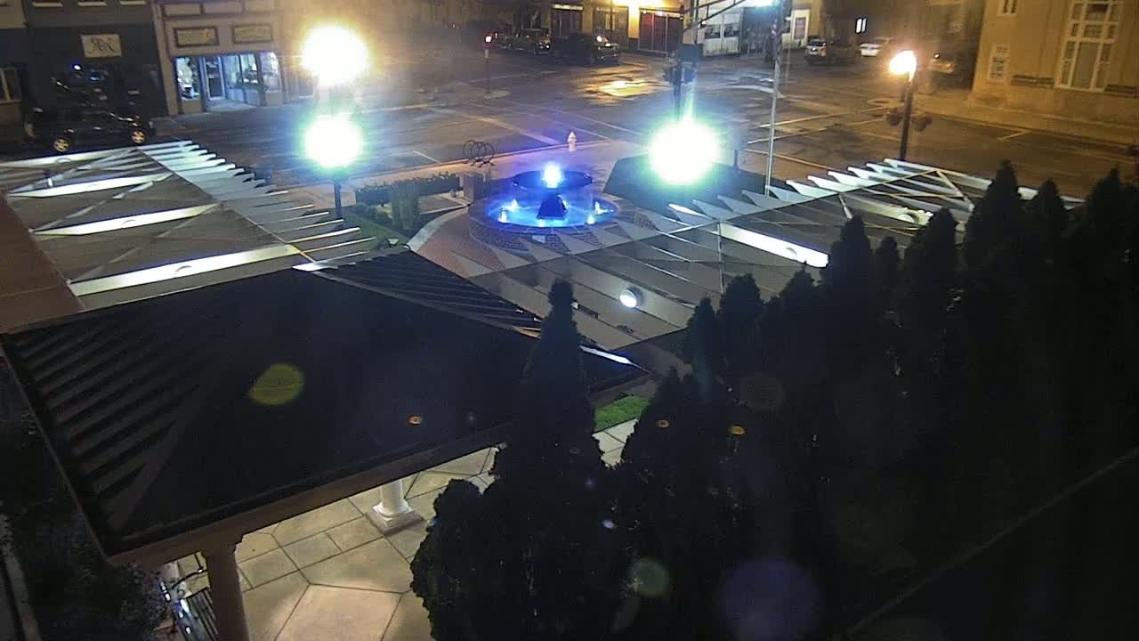 A nighttime, overhead view of a blue illuminated fountain in a plaza surrounded by dark evergreen trees and a building with a dark peaked roof.