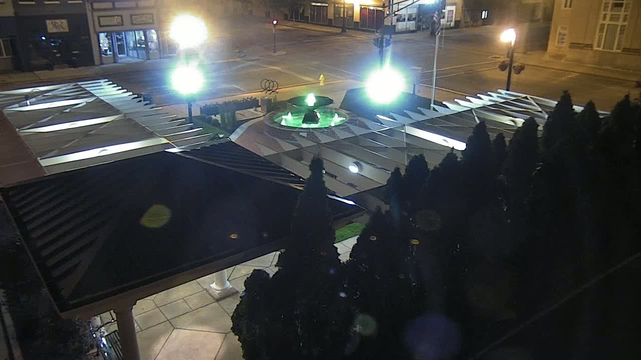 A nighttime, overhead view of a fountain in a town square, surrounded by dark evergreen shrubs and illuminated by streetlights.