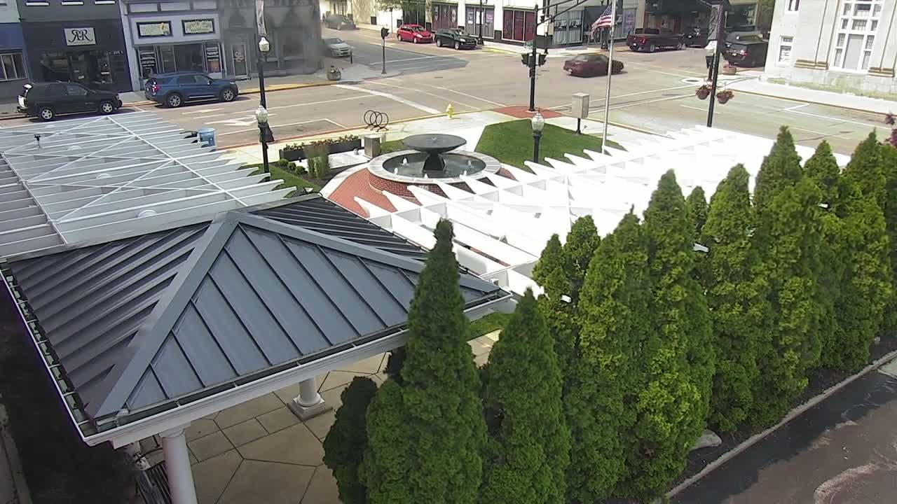 An aerial view shows a town square with a fountain, a white pergola, and a row of evergreen trees on a sunny day.