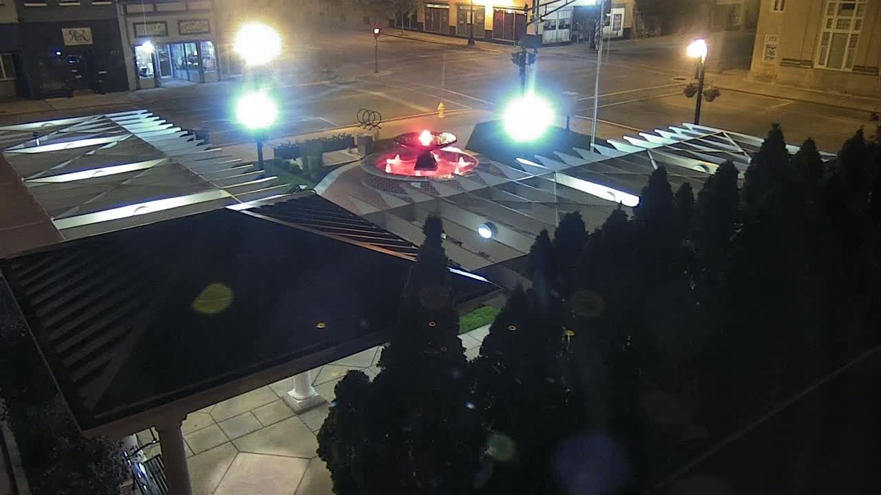 An illuminated nighttime scene shows a red-lit fountain surrounded by evergreen shrubs and streetlights, viewed from above a dark-roofed structure.