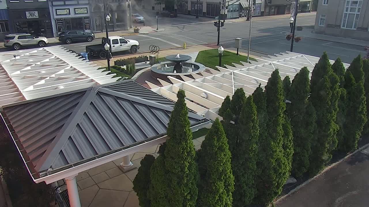 An overhead view of a town square featuring a fountain, a white pergola, and a row of evergreen trees on a sunny day.