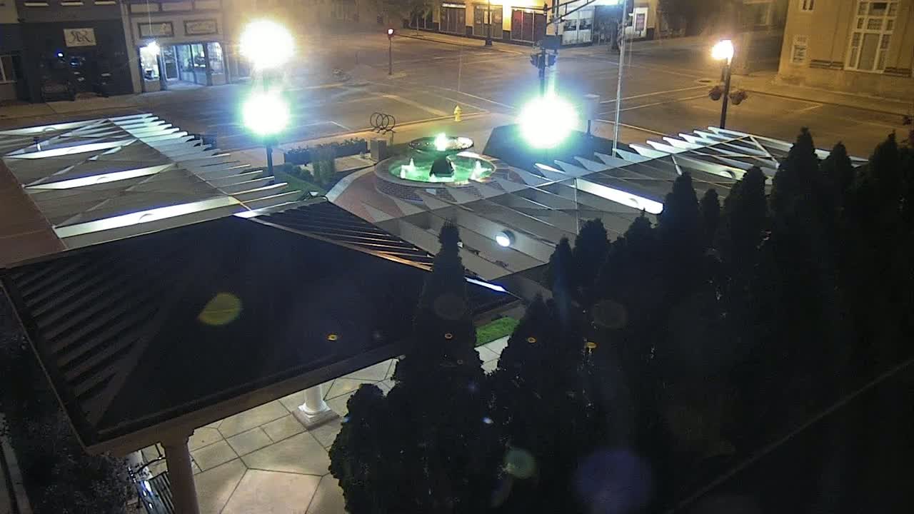 A nighttime aerial view shows a lit fountain in a plaza surrounded by dark evergreen shrubs and a modern, angular building with a covered walkway.