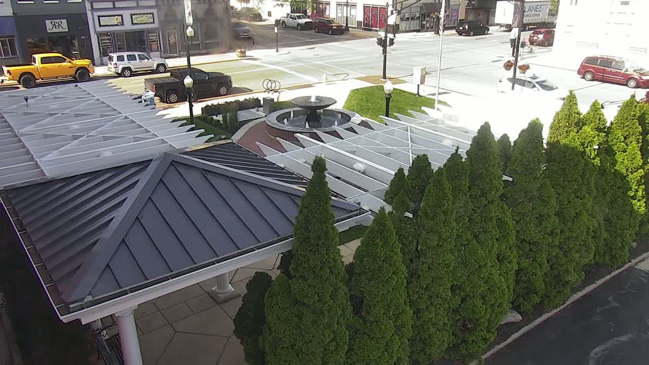 A dark-roofed gazebo sits beside a row of evergreen trees near a fountain in a sunny, paved plaza.